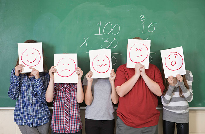 Kids standing in front of a chalk board with a math problem on it. They are holding up drawings of different emotional faces.